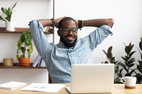 Smiling-Afro-American-businessman-holding-hands-behind-head-sitting-at-office-desk-behind-laptop.